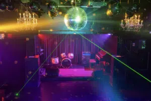 A small stage with a drum set, amplifiers, and a piano is illuminated by colorful lights and a disco ball in a dimly lit venue.